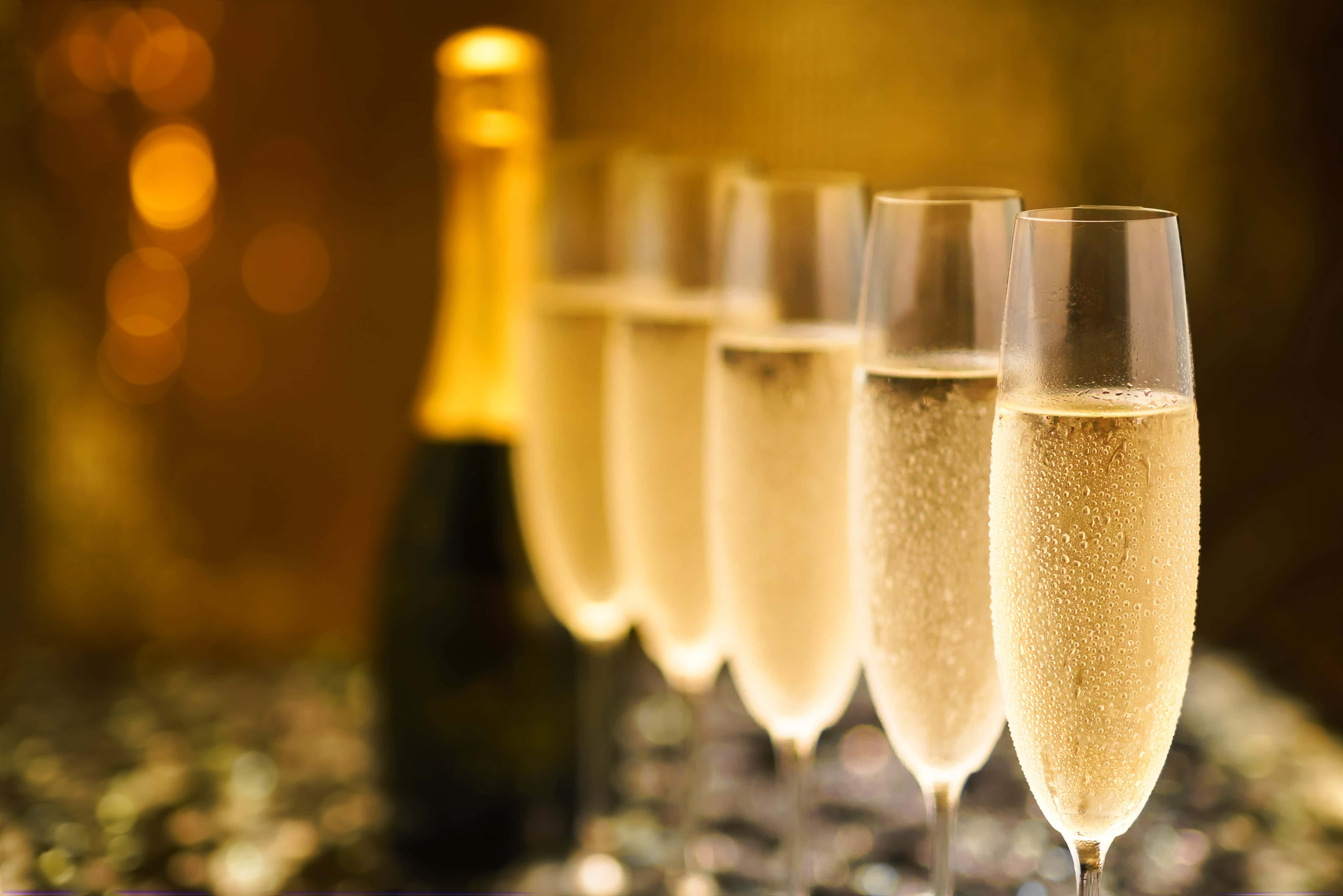 A row of champagne flutes filled with sparkling wine in front of a blurred bottle, set against a warm, golden background with festive bokeh lights.