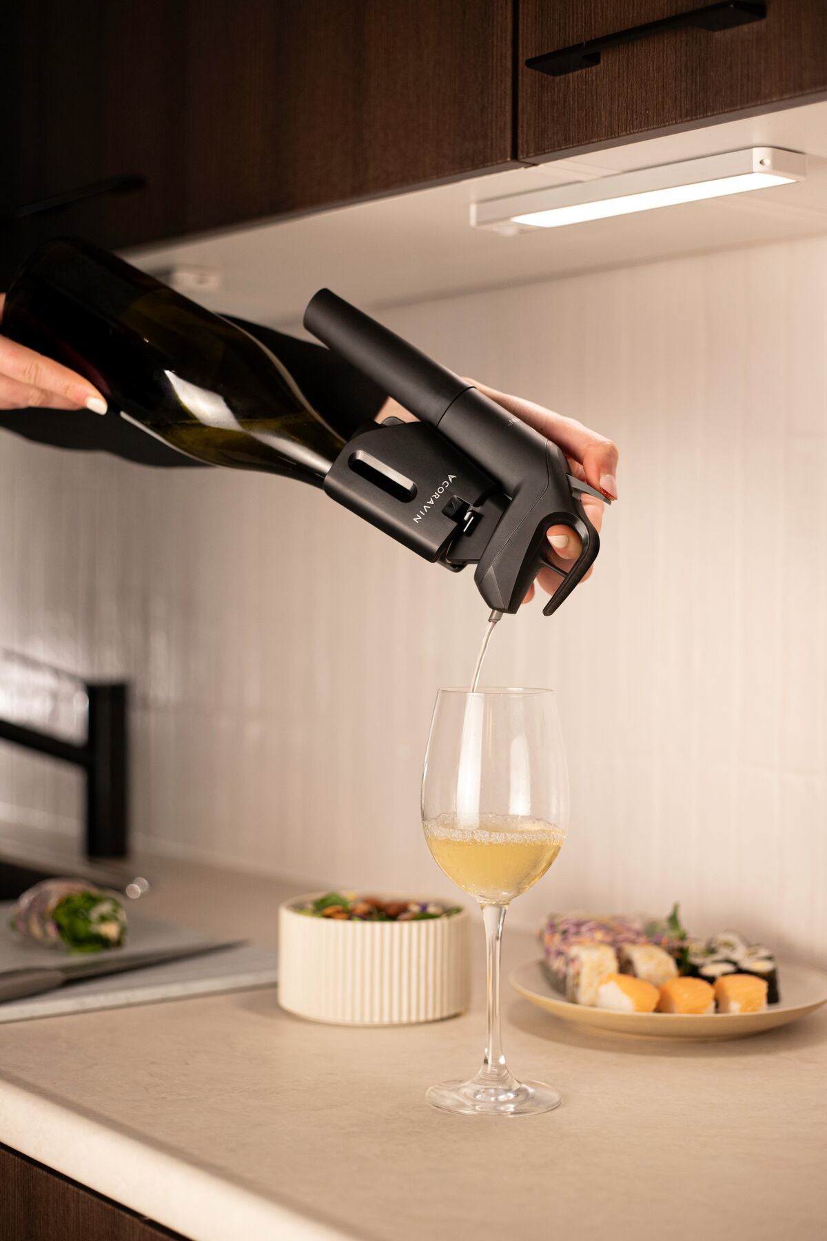 A hand pours sparkling wine from a bottle using a wine pump into a glass on a countertop, with sushi and salad nearby.