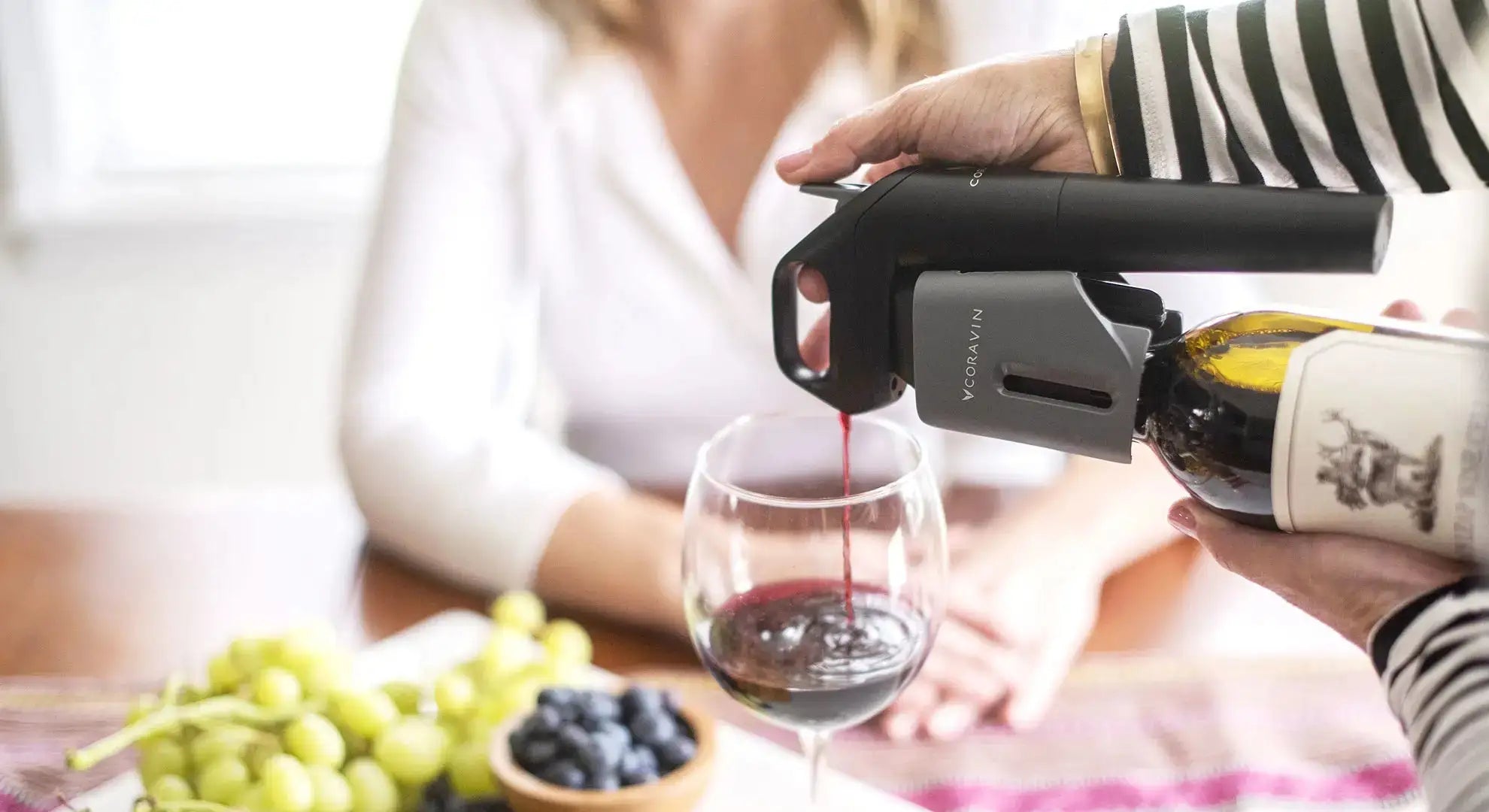 A person uses a Coravin wine opener to pour red wine into a glass, with grapes and blueberries nearby on the table.