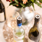 Close-up of wine bottles with Coravin closures, a glass, and a floral arrangement on a sparkling table surface.