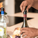 A hand removes the cork from a red wine bottle with two other bottles and a cheese platter in the background.