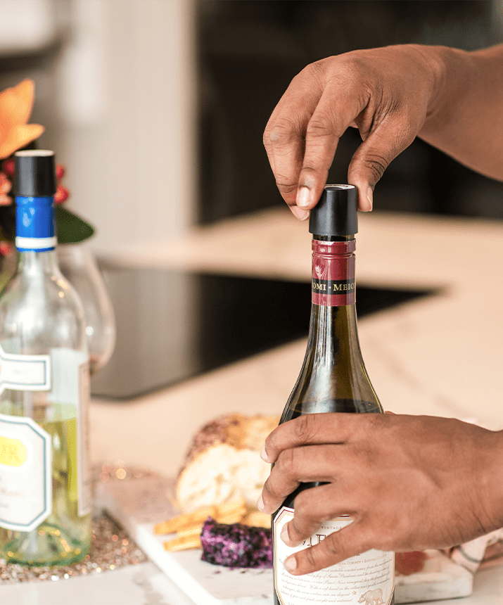 A hand removes the cork from a red wine bottle with two other bottles and a cheese platter in the background.