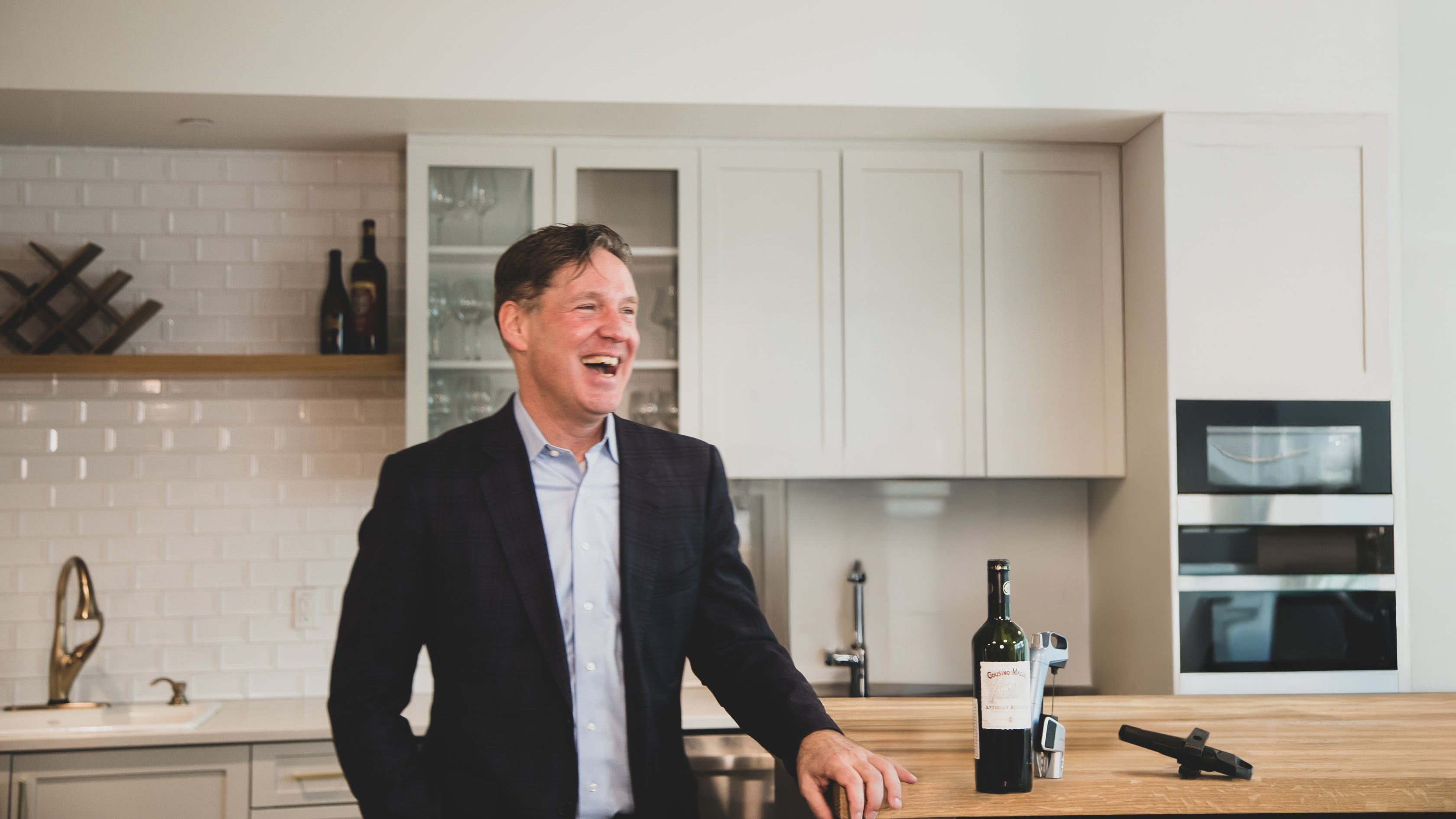 Man in a dark blazer laughs while standing near a bottle of wine and a corkscrew in a modern kitchen setting.