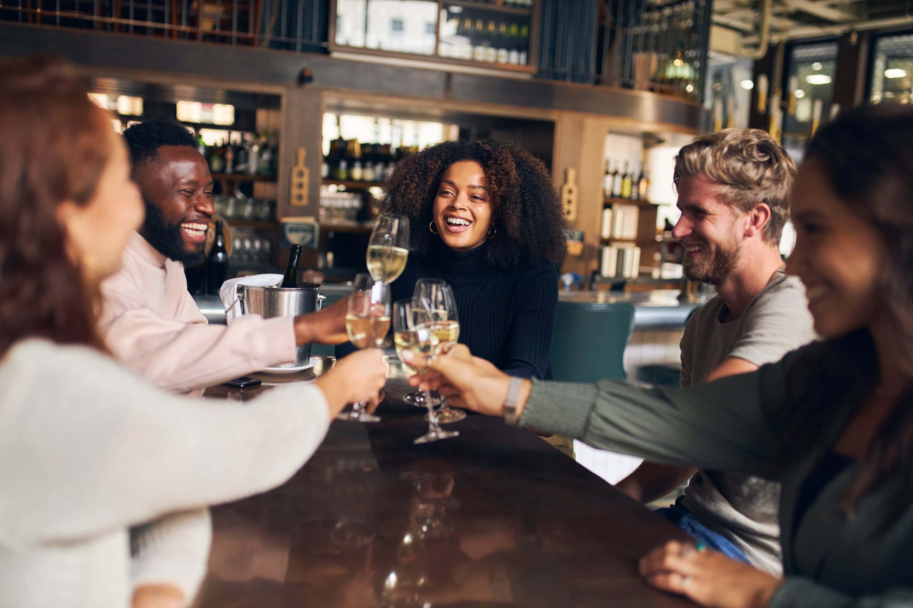 A group of friends gathered around a bar table, smiling and toasting with glasses of white wine in a stylish, modern wine bar setting.