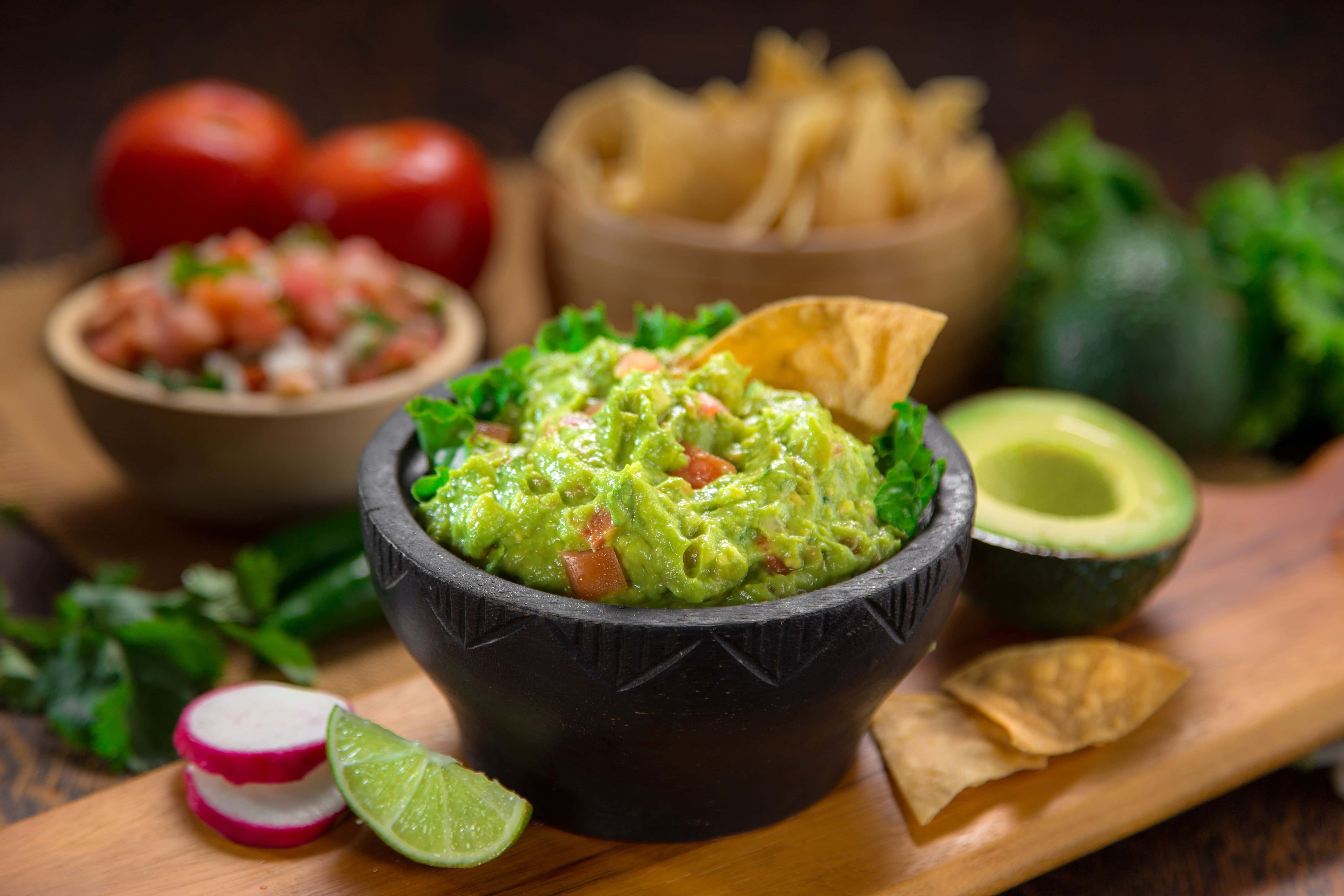 Fresh guacamole served in a traditional black molcajete, garnished with tortilla chips and surrounded by lime slices, radishes, avocado, salsa, and tomatoes.