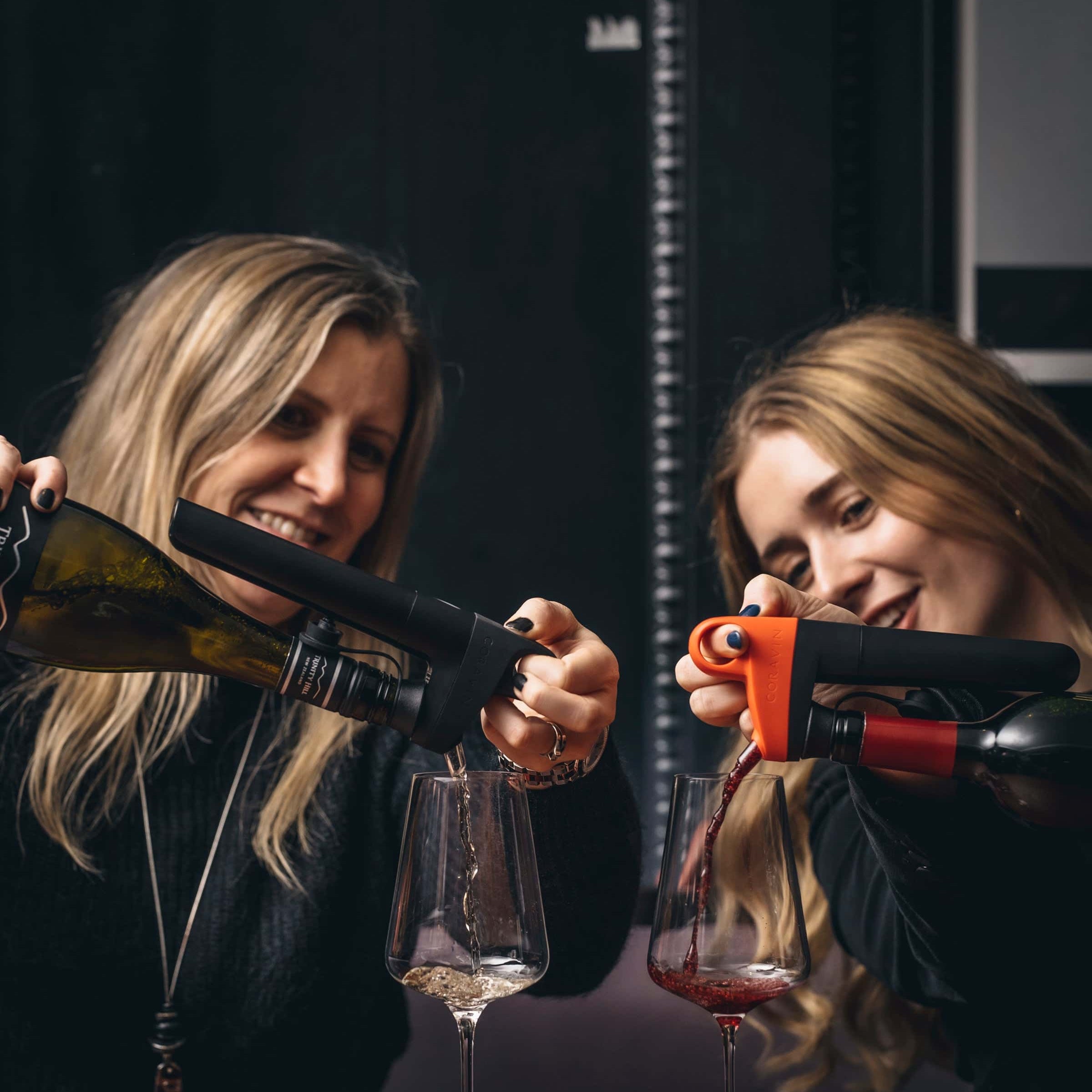 Two women smiling and pouring white and red wine into glasses using Coravin wine preservation systems at a wine tasting session.