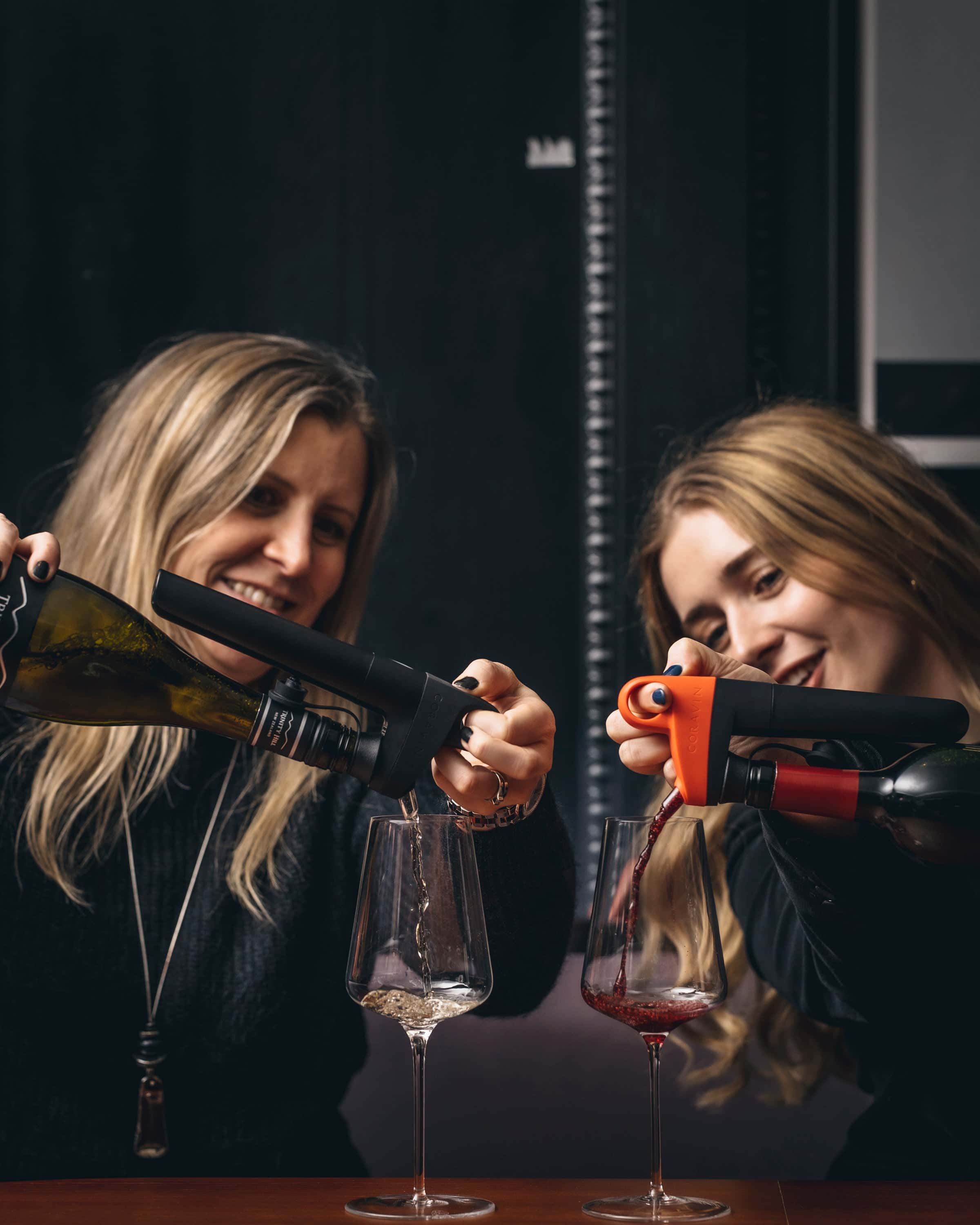 Two women smiling and pouring white and red wine into glasses using Coravin wine preservation systems at a wine tasting session.