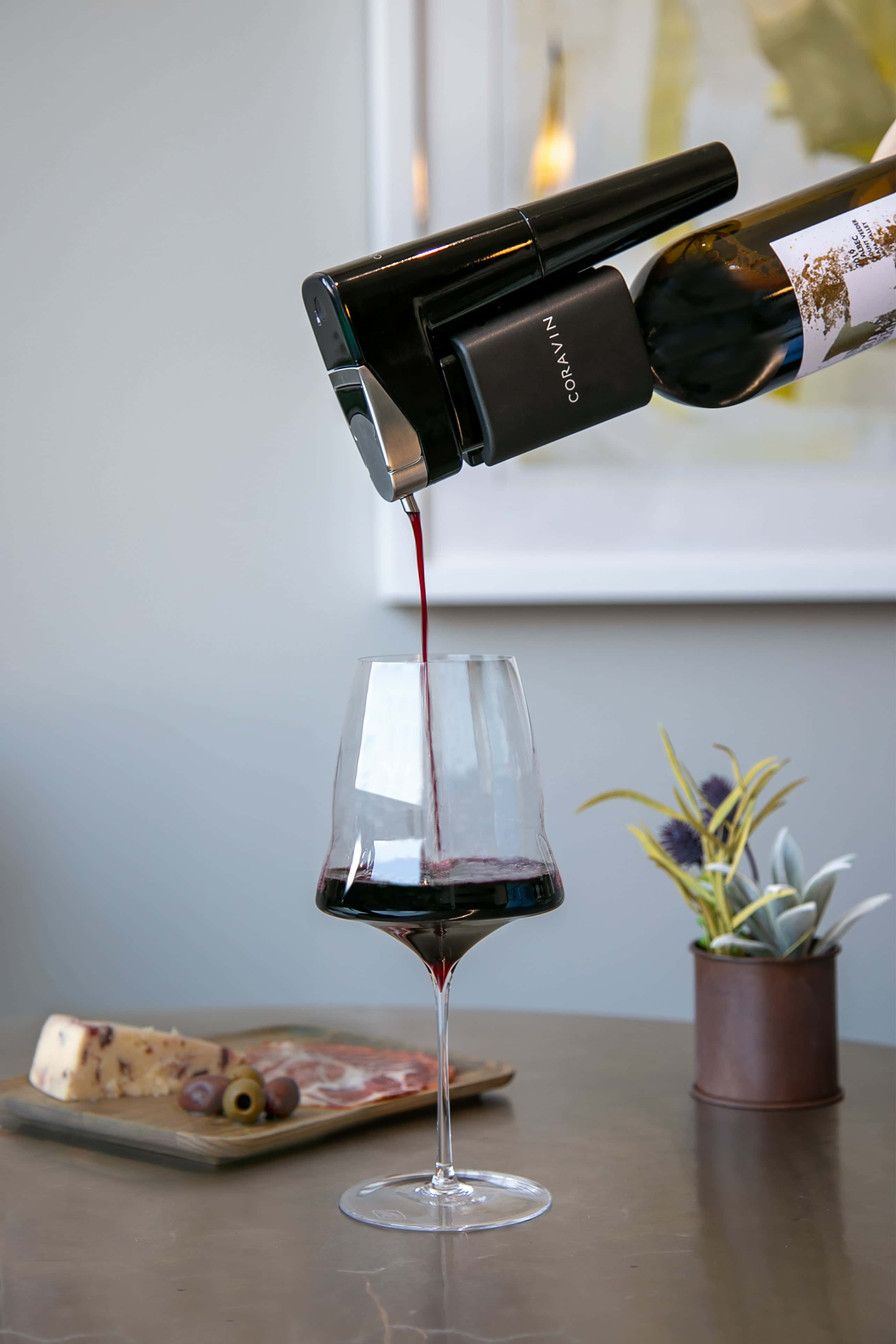 A Coravin wine preservation system pouring red wine into a modern stemmed glass, with a charcuterie board and potted plant in the background on a sleek tabletop.