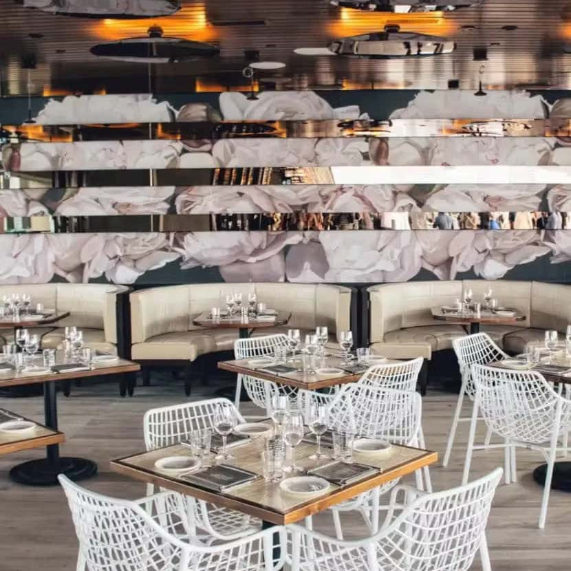 Chic indoor restaurant with modern white lattice chairs, wood-trimmed tables set for dining, curved beige booths, and a large floral mural accented by mirrored horizontal panels on the back wall.