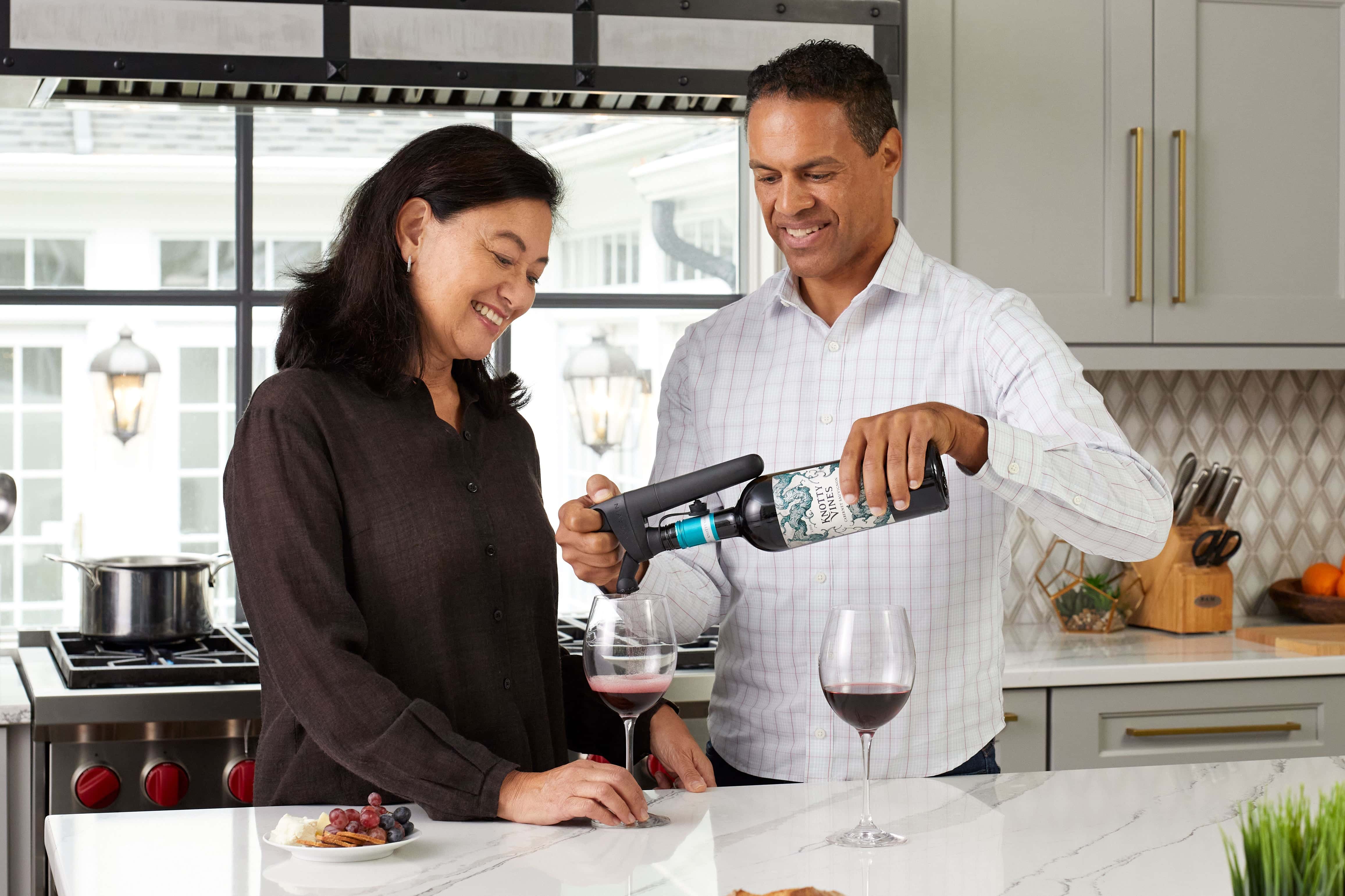 Smiling couple pouring red wine with a Coravin Pivot wine preservation system in a modern kitchen setting.