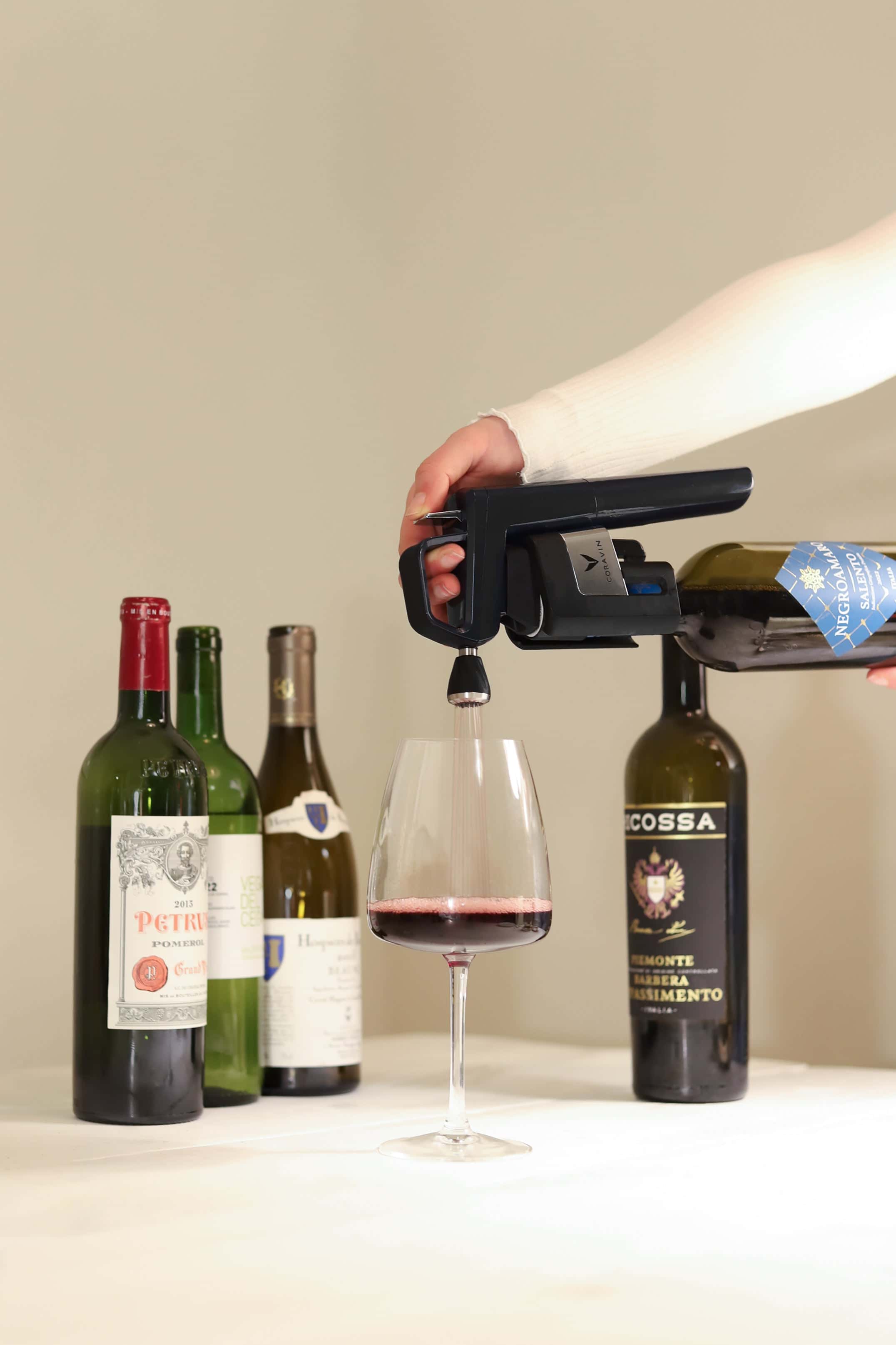 Hand using a Coravin wine preservation system to pour red wine into a glass, with several wine bottles in the background on a white tablecloth.