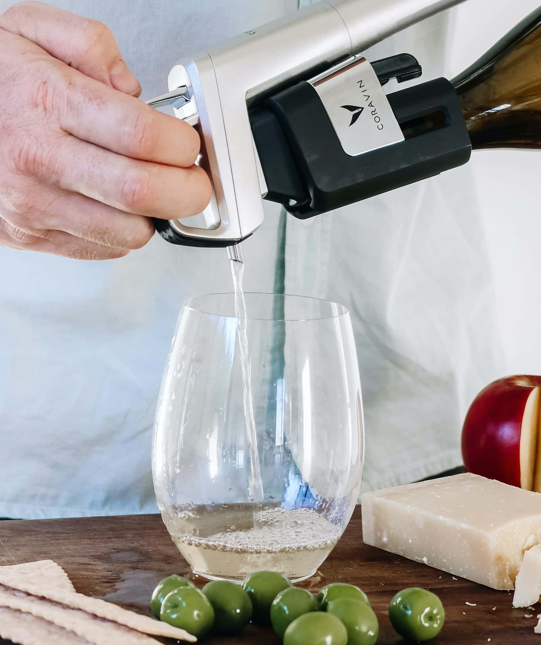 Close-up of a hand pouring white wine using a Coravin wine preservation system into a stemless glass, with green olives, crackers, a wedge of cheese, and a sliced apple on a wooden surface.