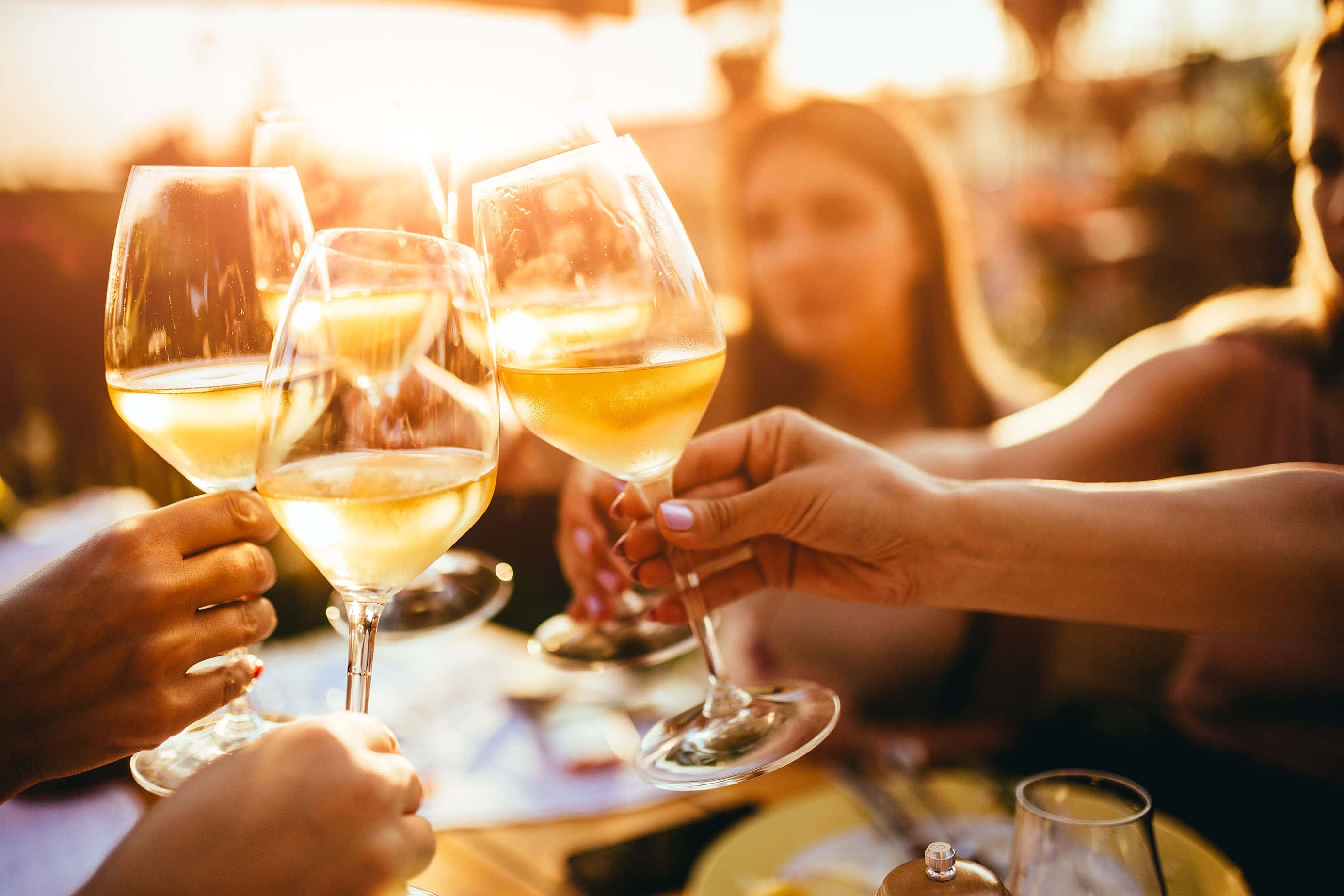 Group of friends toasting with glasses of white wine at an outdoor gathering during golden hour, with sunlight illuminating the drinks and warm, cheerful atmosphere.