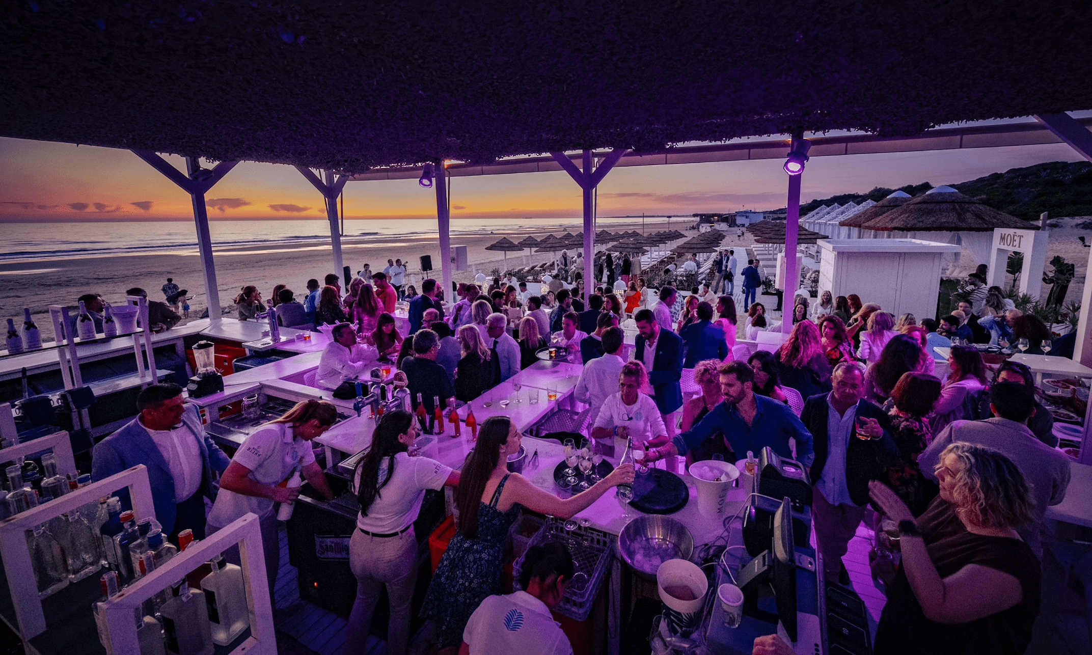Crowded beach bar at sunset with people enjoying cocktails and wine under purple lights.