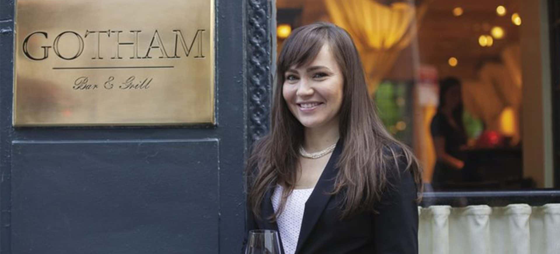 Woman smiling in front of Gotham Bar & Grill sign, dressed in a black blazer and holding a wine glass, standing outside the restaurant.