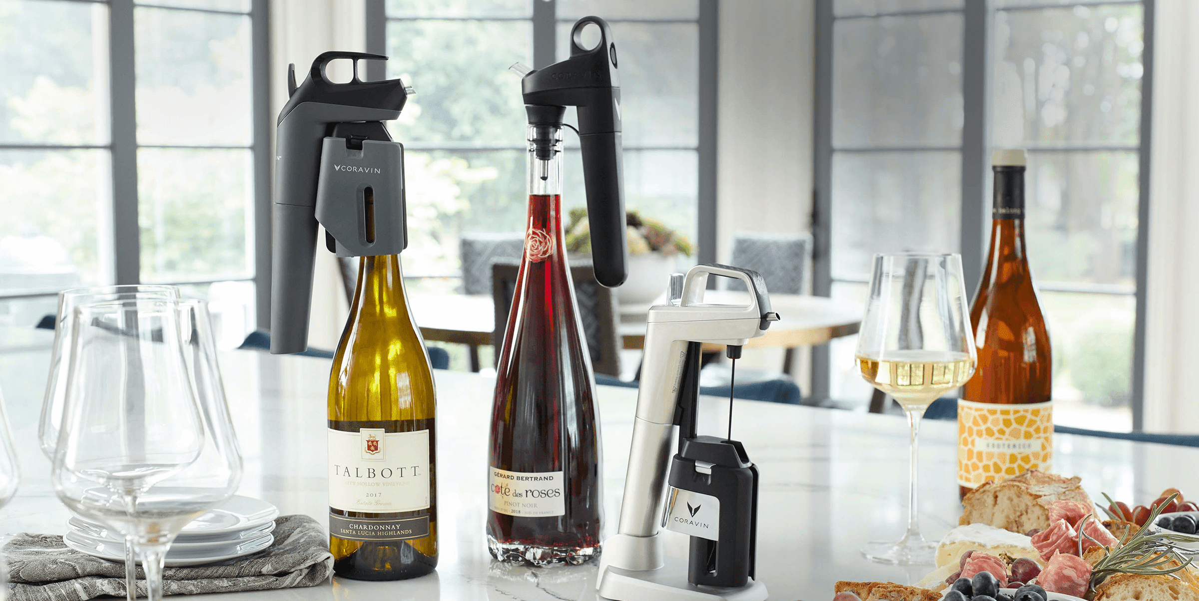 A selection of Coravin wine preservation systems displayed on a kitchen counter with various bottles of wine, wine glasses, and a charcuterie board in a bright, modern dining space.