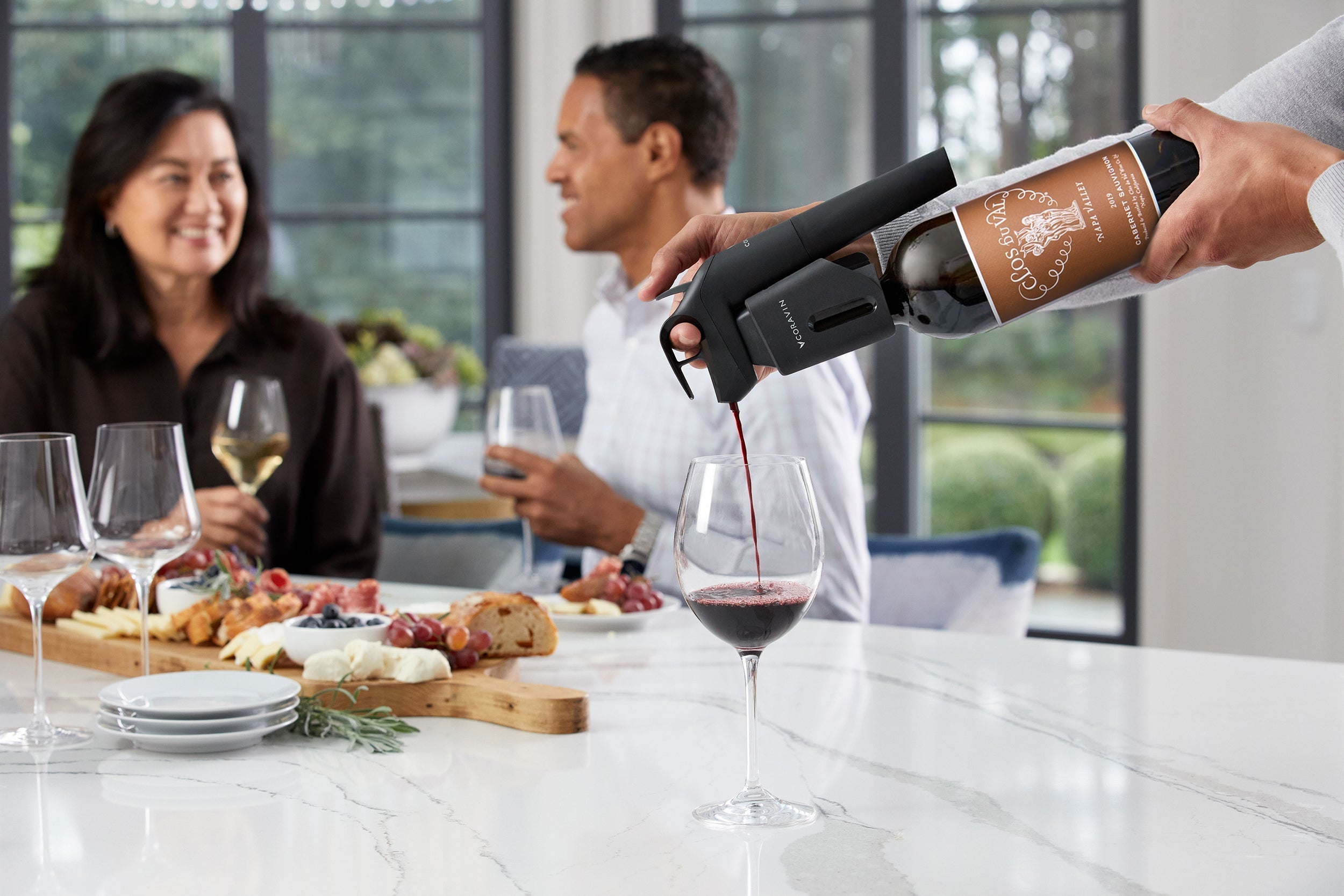 Pouring red wine with the Coravin Timeless wine preservation system at a social gathering, with guests enjoying wine and a charcuterie board on a marble countertop.