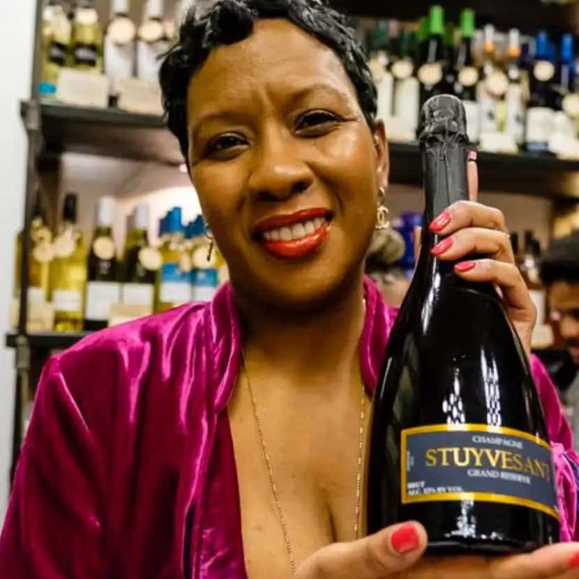 Smiling woman in a pink velvet outfit holding a bottle of Stuyvesant Champagne in a wine shop, with shelves of assorted wine bottles in the background.