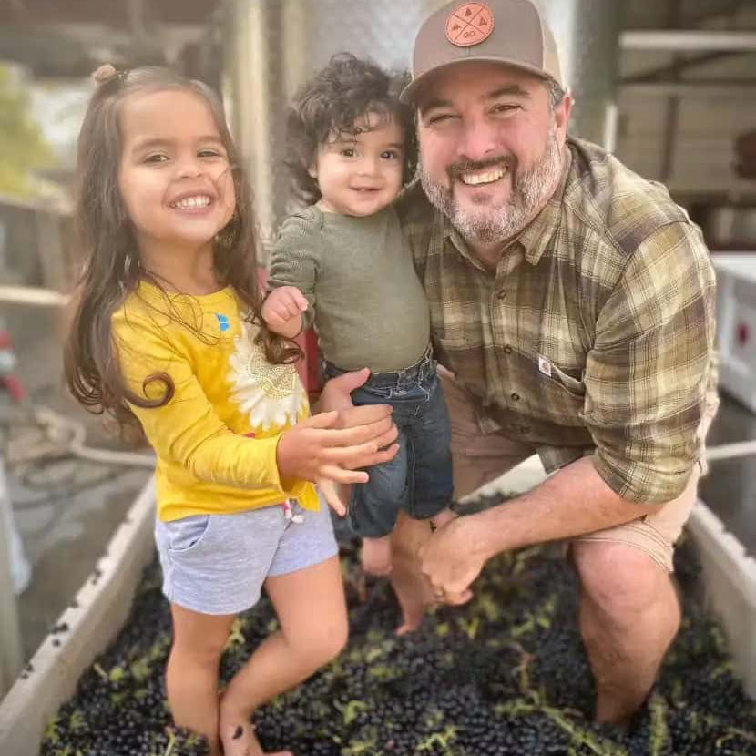 Smiling man with two young children standing barefoot in a large container filled with harvested grapes during winemaking.