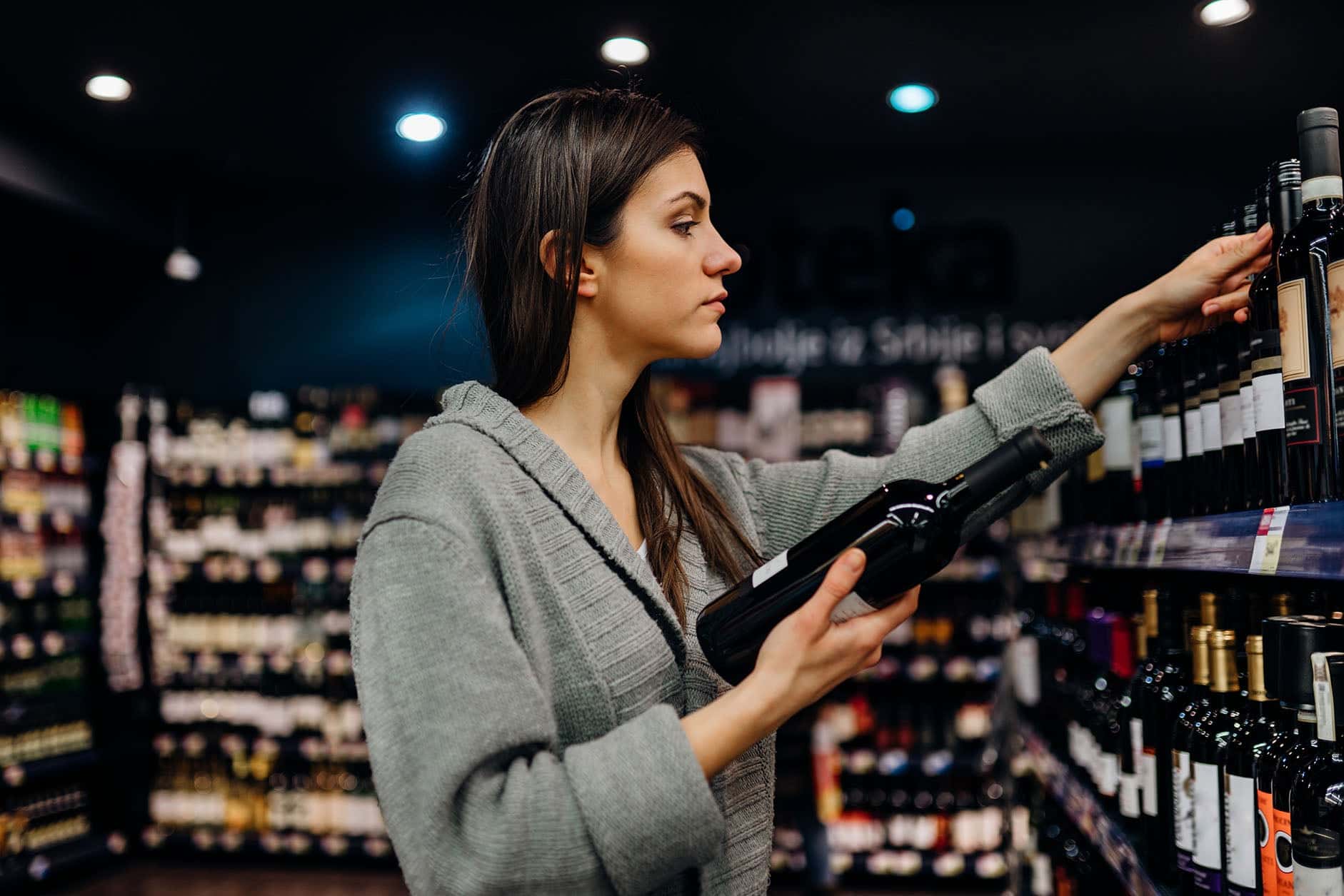 Woman browsing and selecting bottles of wine from shelves in a liquor store or wine shop.