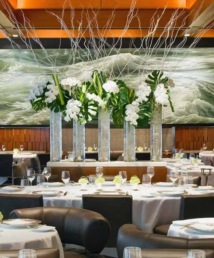 Elegant fine dining restaurant interior with round tables set with white tablecloths, glassware, and floral centerpieces, featuring tall glass vases with white orchids and large green leaves in the center of the room.