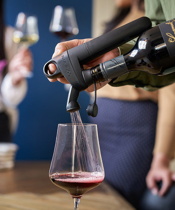 A Coravin wine pouring system dispenses red wine into a glass, with blurred glasses of white wine in the background.