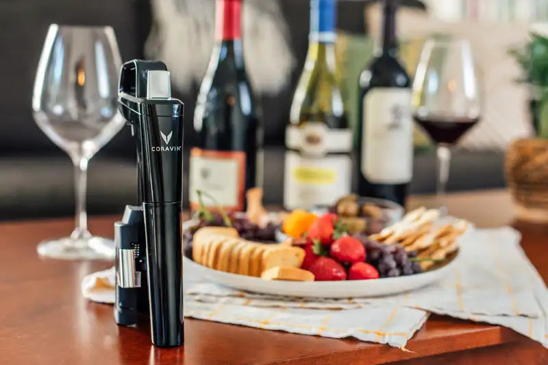 Coravin wine opener beside a plate of fruits, crackers, and wine bottles, with glasses in a cozy setting.