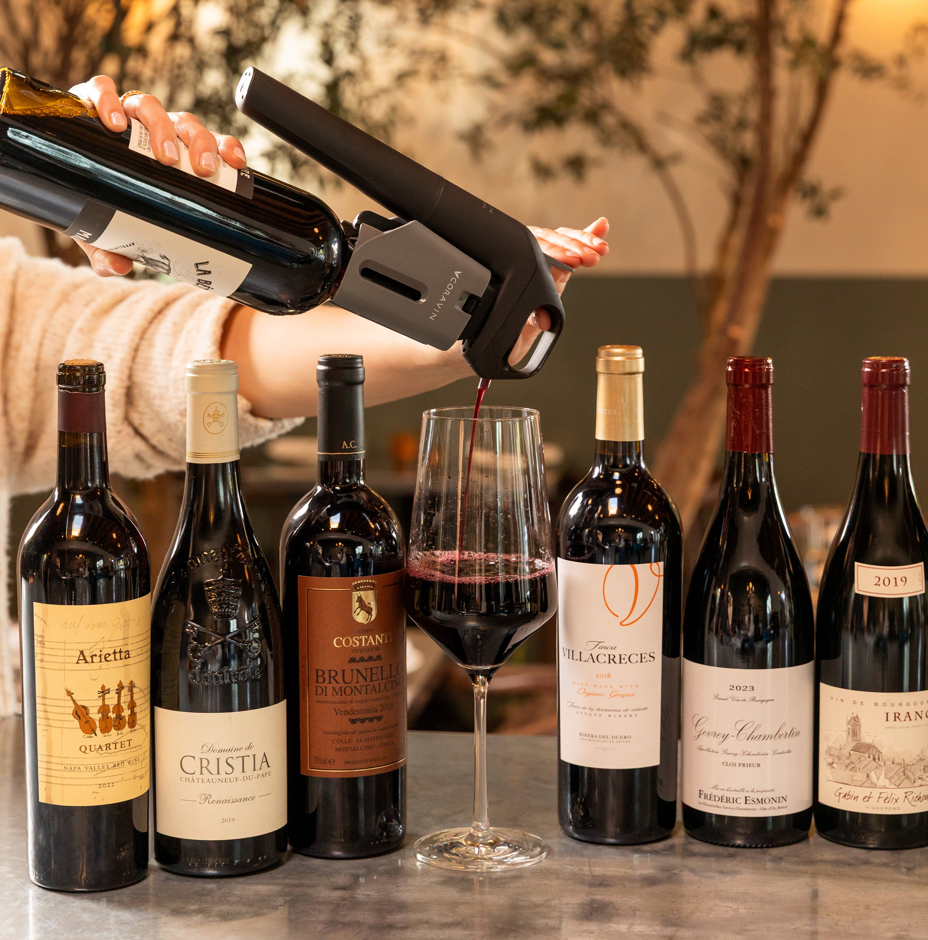 Pouring red wine using a Coravin Timeless Six+™ wine by-the-glass system at a wine bar, surrounded by premium red wine bottles including Brunello, Châteauneuf-du-Pape, and Napa Valley wines.